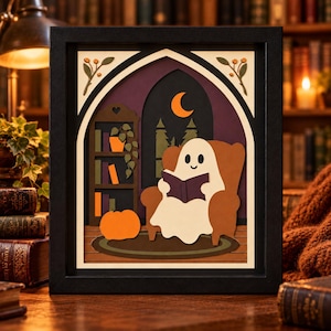 May include: A layered paper art shadow box in a black frame featuring a white ghost sitting in a brown armchair reading a purple book. The scene includes a bookshelf, a pumpkin, a crescent moon, and a forest silhouette against a dark purple background.