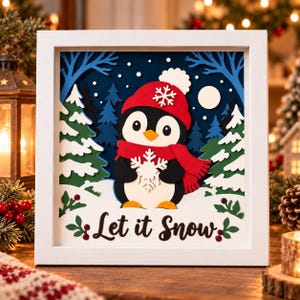 May include: A white framed shadow box featuring a layered paper cutout of a penguin wearing a red hat and scarf, holding a white snowflake. The scene includes green and blue pine trees, a white moon, and the text Let it Snow in black.