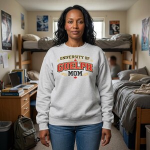 May include: A light grey sweatshirt with the text "UNIVERSITY OF GUELPH MOM" in red, black, and gold. The sweatshirt has a crew neck and long sleeves. The woman is wearing blue jeans.