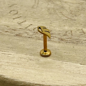 May include: A gold-colored lip piercing stud with a leaf design. The stud features a flat base, a slender post, and a leaf-shaped top with two cut-out details. The jewelry is set against a light-colored wooden surface.