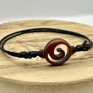 May include: A minimalist bracelet featuring a dark brown wooden spiral charm on a black cord. The bracelet is knotted on each side of the charm. The bracelet is displayed on a wooden surface.