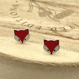 May include: A pair of fox-shaped stud earrings. Each earring features a red fox head with white and black accents. The earrings are set against a light brown wooden surface with the words "LIMITED EDITION" in a circular pattern.