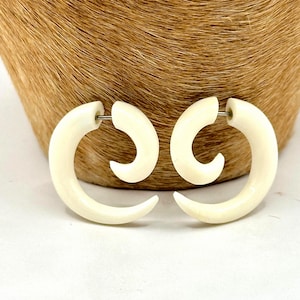 May include: A pair of off-white spiral-shaped earrings featuring a curved, tapered design that mimics the appearance of a tusk. Each earring has a thin metal post extending from the inner curve, designed to pass through a pierced earlobe.