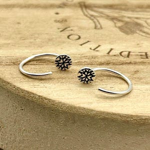 May include: A pair of silver open hoop earrings featuring a small, circular flat end piece on each. Each circular end is engraved with a black, star-like geometric pattern. The earrings have a smooth, polished finish and a curved, open-ended design.
