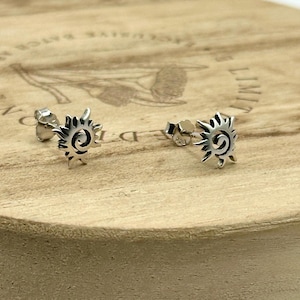 May include: A pair of silver-colored stud earrings. Each earring features a sunburst design with a central spiral. The earrings are displayed on a light brown wooden surface.