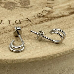 May include: A pair of silver stud earrings with a double-hoop design. The earrings are displayed on a light-colored wooden surface. The earrings have a post and butterfly backing.