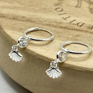 May include: A pair of silver hoop earrings with a clear gemstone and a small seashell charm. The earrings are displayed on a wooden surface.