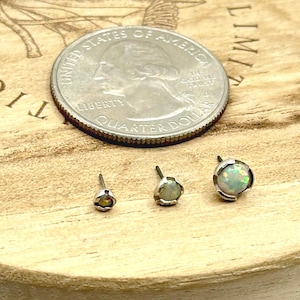 May include: Three opal stud earrings of varying sizes are displayed next to a quarter. The earrings have a silver setting and a colorful, iridescent opal stone. The earrings are on a wooden surface.