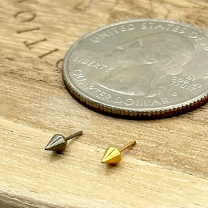 May include: Two small, spiked earrings, one gold and one silver, are displayed on a wooden surface next to a quarter. The earrings have a simple, pointed design. The quarter is silver and shows the profile of George Washington.