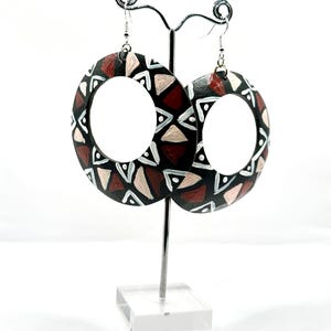 May include: A pair of large, circular earrings with a black base, adorned with geometric patterns in white, tan, and burgundy. The earrings are displayed on a silver metal stand against a white backdrop, showcasing their bold design and craftsmanship.
