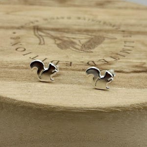 May include: A pair of silver-colored squirrel stud earrings. The earrings are small, with a detailed squirrel design. They are displayed on a round, light-colored wooden surface.