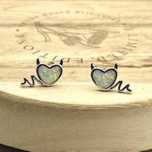 May include: A pair of silver-toned stud earrings. Each earring features a heart-shaped opal stone with a devil design, including horns and a curved tail. The earrings are set on a wooden surface.