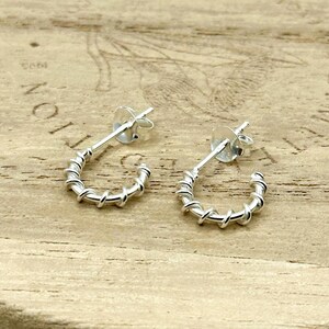 May include: A pair of silver hoop earrings featuring a twisted wire design. Each earring is a small, semi-circular hoop with a post back and a circular earring back closure. The silver metal has a polished finish that reflects light across the surface.