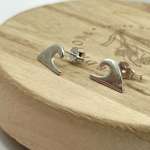 May include: A pair of silver wave-shaped stud earrings. The earrings are displayed on a light brown wooden surface. The earrings have a polished finish and a post backing.