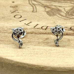 May include: A pair of silver fox stud earrings. Each earring features a fox head and curved tail, both embellished with small, clear gemstones. The earrings are set against a light brown wooden surface.