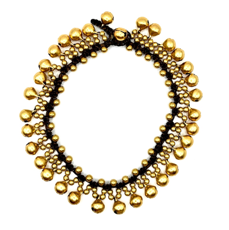 K&ouml;nnte beinhalten: Ein kreisf&ouml;rmiges Fu&szlig;kettchen mit einer Basis aus dunkelbrauner Kordel, die mit zahlreichen kleinen, metallisch goldfarbenen, kugelf&ouml;rmigen Gl&ouml;ckchen verziert ist. Die Gl&ouml;ckchen sind in einem sich wiederholenden Muster entlang der Kordel angeordnet.