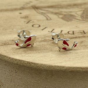 May include: A pair of silver stud earrings shaped like koi fish, with red enamel markings. The earrings are displayed on a wooden surface, showcasing their design and detail. These earrings are a unique accessory.