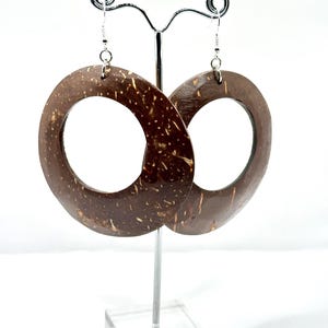 May include: A pair of large, open-circle earrings made from dark brown coconut shell. The earrings are suspended from silver-colored hooks and displayed on a silver earring stand against a white background. The earrings have a natural, textured appearance.