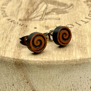 May include: A pair of wooden stud earrings with a spiral design. The earrings have a black border and a brown spiral center. The earrings are on a wooden surface.