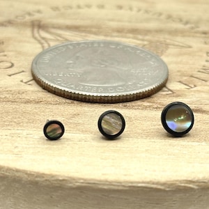May include: Three black-rimmed, iridescent stud earrings of varying sizes are displayed next to a quarter coin on a wooden surface. The earrings have a circular design with a shimmering, multi-colored center.