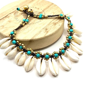 May include: An anklet featuring a dark brown cord woven with small turquoise beads and metallic gold-toned beads arranged in a floral pattern. The piece is adorned with a series of white cowrie shells hanging along the length of the cord.