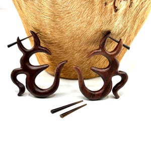 May include: Pair of dark brown wooden earrings with an antler-like design. The earrings have a curved, organic shape with multiple points and a straight post. Two additional wooden posts are also visible.