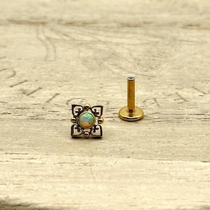 May include: A gold-colored, decorative piercing stud with a square, filigree design and a central opal-like stone. Next to it is a gold-colored post with a flat base. The items are on a light-colored wooden surface.
