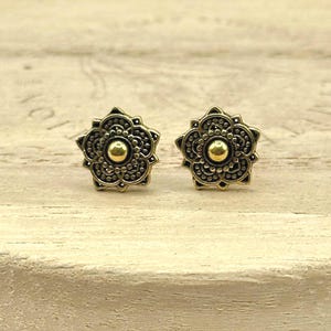 May include: A pair of gold-colored stud earrings. Each earring features a floral design with a central gold-colored sphere. The earrings are set against a light-colored wooden surface.