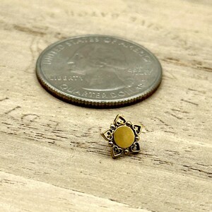 May include: A small, gold-colored star-shaped stud earring with a central circular design and heart-shaped cutouts. A quarter coin is next to the earring for size comparison. The earring is on a wooden surface.