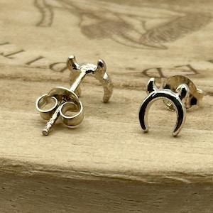 May include: A pair of silver-colored stud earrings in the shape of a crescent moon with two small horns. The earrings are displayed on a wooden surface.