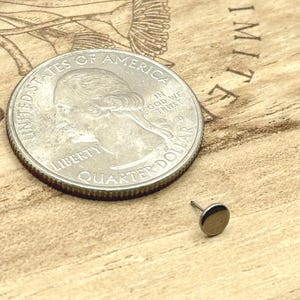 May include: A small, round, gold-colored stud earring next to a U.S. quarter coin. The earring is on a light brown wooden surface with etched designs and the word "IMITATE". The quarter is silver with the words "LIBERTY" and "QUARTER DOLLAR".