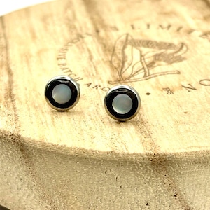 May include: A pair of small stud earrings. Each earring features a black border surrounding a white, circular center. The earrings are set on a light brown wooden surface with etched text and a graphic.