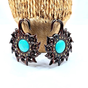 May include: Pair of dark brown wooden earrings with a turquoise center stone and gold-colored accents. The earrings have a sunburst design with intricate carved details. The background includes a woven basket.