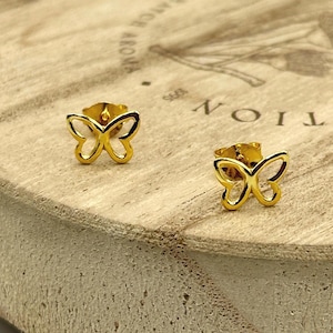 May include: A pair of gold-colored butterfly stud earrings. The earrings are displayed on a light brown wooden surface. The butterfly design is open, with the shape of the butterfly wings clearly visible.