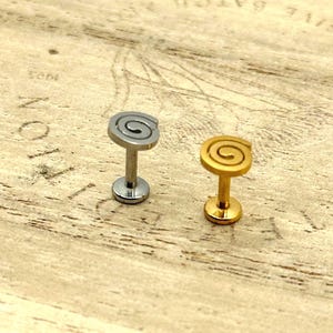 May include: Two spiral-shaped labret studs. One is silver-colored, the other is gold-colored. The studs have a flat base and a post. The spiral design is on the top of the stud. The studs are on a light brown wooden surface.