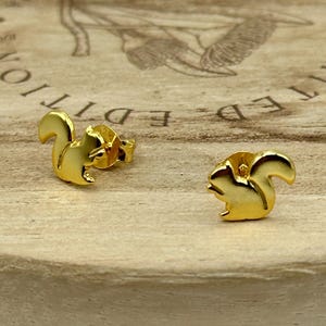 May include: A pair of gold-colored squirrel stud earrings. The earrings are small, detailed, and have a shiny finish. They are displayed on a light brown wooden surface with the words "LIMITED EDITION" partially visible in the background.