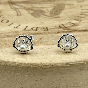 May include: A pair of sterling silver seashell stud earrings with a small pearl in the center of each shell. The earrings are set against a light brown wooden surface.