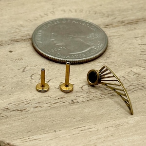 May include: Close-up of three gold-colored body jewelry pieces and a quarter. Two pieces are small, with a flat base and a post. The third piece has a fan-like design with a dark center. The items are on a wooden surface.