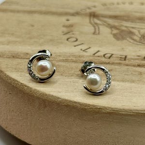 May include: A pair of silver-toned stud earrings. Each earring features a white pearl cradled within a crescent-shaped design, embellished with small, clear crystals. The earrings are displayed on a wooden surface.