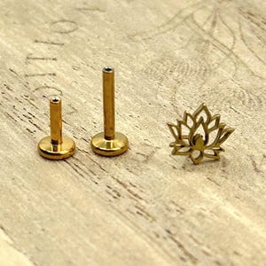 May include: Gold-colored body jewelry, including two labret studs and a lotus flower stud. The labret studs have a flat disc base and a post. The lotus stud is a cutout design. All pieces are displayed on a light wood surface.