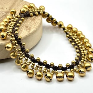 K&ouml;nnte beinhalten: Ein Fu&szlig;kettchen aus dunkelbrauner Kordel mit mehreren Reihen kleiner, runder, goldfarbener Metallgl&ouml;ckchen. Die Gl&ouml;ckchen sind in einem dichten, geschichteten Muster an der geflochtenen Kordel befestigt und bilden ein durchgehendes, gl&auml;nzendes Design.