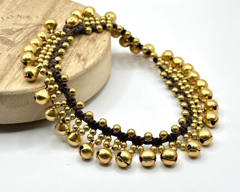 K&ouml;nnte beinhalten: Ein Fu&szlig;kettchen aus dunkelbrauner Kordel mit mehreren Reihen kleiner, runder, goldfarbener Metallgl&ouml;ckchen. Die Gl&ouml;ckchen sind in einem dichten, geschichteten Muster an der geflochtenen Kordel befestigt und bilden ein durchgehendes, gl&auml;nzendes Design.