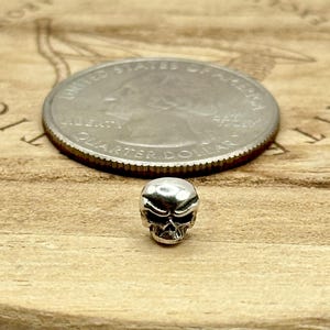 May include: A small, silver skull bead is positioned in front of a U.S. quarter coin on a wooden surface. The skull has detailed facial features. The quarter coin is silver and shows the profile of George Washington.