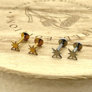 May include: Four star-shaped stud earrings. Two are gold-toned, and two are silver-toned. The earrings are displayed on a light-colored wooden surface with etched text and a graphic.