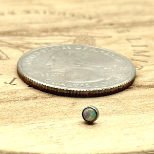 May include: A small, iridescent opal stud earring next to a U.S. quarter coin on a wooden surface. The earring is a small, round shape with a subtle, colorful sheen. The quarter is silver with detailed engravings.