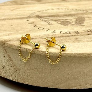 May include: A pair of gold-colored stud earrings with a small ball and a delicate chain. The earrings are displayed on a light brown wooden surface. The earrings are a simple and elegant jewelry item.