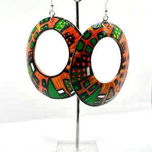 May include: Oval-shaped earrings with a colorful, geometric design. The earrings feature a pattern of orange, green, black, and white shapes. The earrings are displayed on a silver earring stand against a white background.