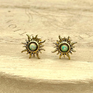 May include: A pair of gold-colored sun-shaped stud earrings. Each earring features a central, round, opalescent stone surrounded by spiky rays. The earrings are displayed on a light-colored, textured surface.