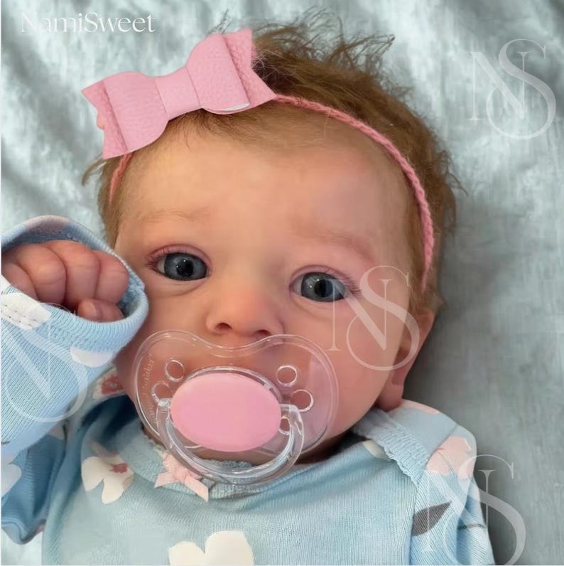 May include: A baby with blue eyes wearing a pink bow headband and a light blue onesie with a floral pattern. The baby has a pink pacifier in its mouth. The image has the text "NamiSweet" in the top left corner.
