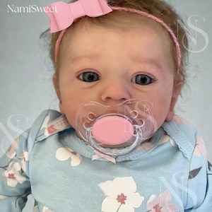 May include: A baby doll with blue eyes, wearing a light blue floral onesie and a pink bow headband. The doll has a pink pacifier in its mouth. The text "NamiSweet" is visible in the upper left corner.